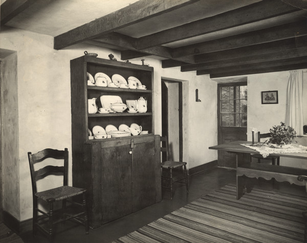 Interior view of the dining room at Trelawny House. A sideboard displaying china is in the center against a wall, flanked by two chairs. The dining table is near an exterior door and window on the right.