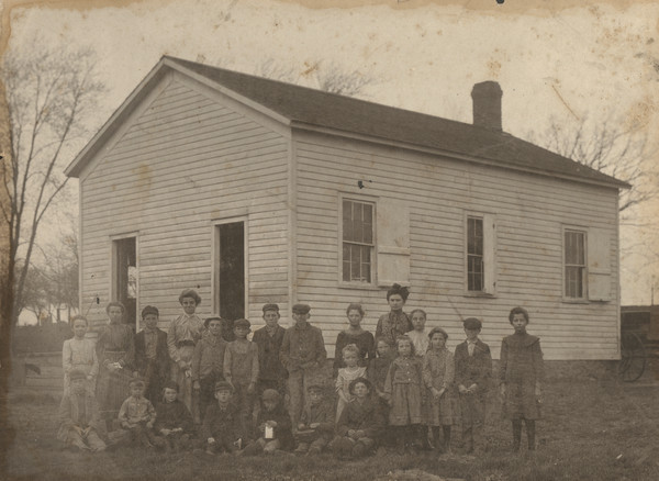 Group portrait of a school class outdoors in front of their one-room schoolhouse.