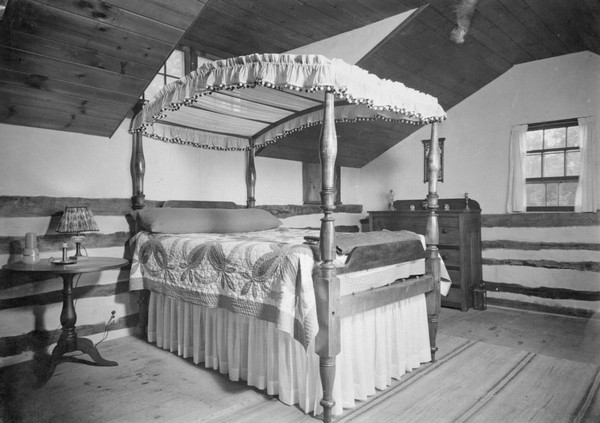 Bedroom in Polperro House with a canopy bed set into a dormer with a window. A quilt is on the bed, a bedside table on the left, and a dresser on the far wall near another window.
