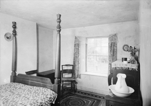 Bedroom in Trelawny House. Furniture includes a 4-poster bed, two chairs, a chest, a dresser, and a wash basin and ewer on a stand.