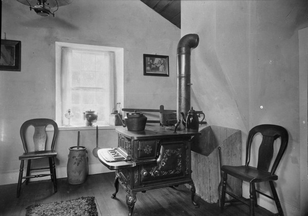 Kitchen at Polperro House. Near the window is a chair and a butter churn. In the center is a woodburning stove, with a firewood box behind it against the wall.