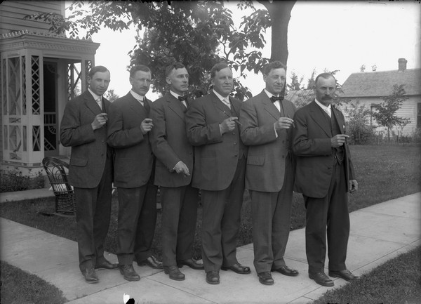 Outdoor group portrait of the six Schmitt brothers, Sherwin Gillett's brothers-in-law. In birth order they are: Charles (b. 1868), Alfred (b. 1870), Louis (b. 1873), Christoph (b. 1876), Otto (b. 1879), and Herbert (b. 1882). They are standing on a sidewalk in front of a house and they are all holding cigars.