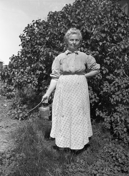 Emma Gillett, Sherwin Gillett's mother, standing outdoors next to a hedge. She is holding a pail and a stick in her right hand.
