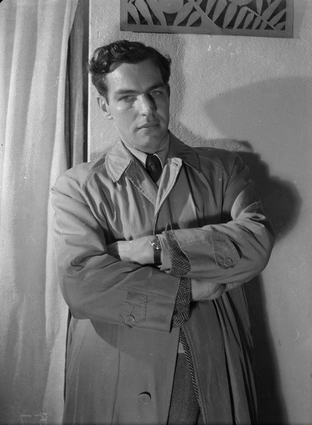 Indoor portrait of Lorin Gillette wearing a coat and leaning against a wall with his arms crossed.