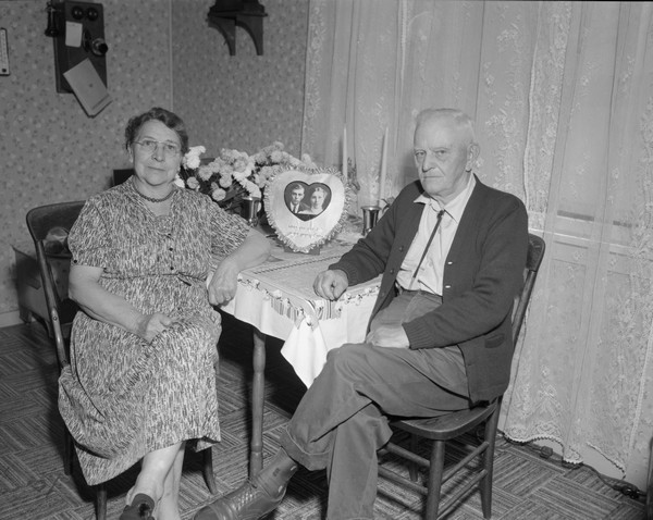 Sherwin and Emma Gillett at their 50th Wedding Anniversary, seated in front of a photograph of them in a heart-shaped frame with the caption: "When you and I were young, Emma."
