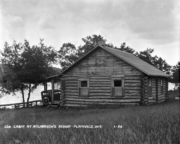View across lawn of long grass towards a log cabin next to the Wisconsin River. An automobile is parked on the left near the porch.