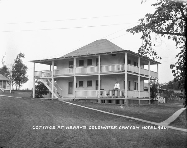 View across lawn of Berry's Coldwater Canyon Hotel. Sidewalks lead to a two-story building with wrap-around porches and balcony. There are exterior stairs and a man is standing on the porch. Other buildings are in the background on the left and right.