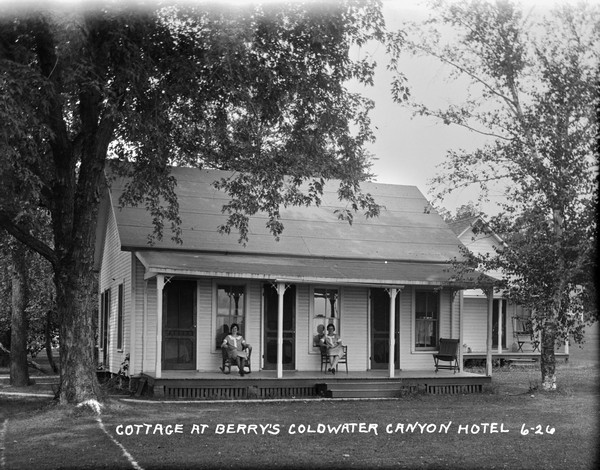 Exterior view of a cottage with three entrances. Two hotel maids are seated on the porch. There is another cottage on the right.