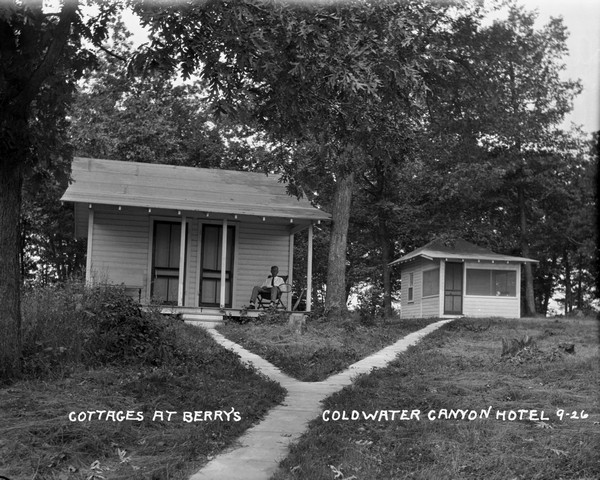 View up slope towards two small cottages at the Coldwater Canyon Hotel. A man with a tennis racket is seated on the porch of the cottage on the left.