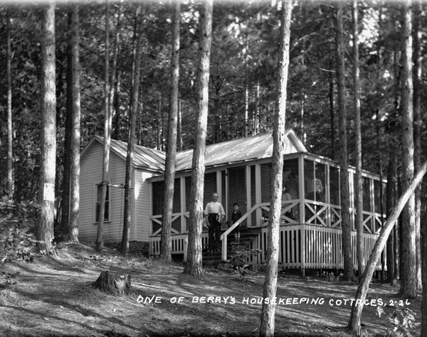 Group of people posed on the steps and porch of a cottage. The cottage has a wrap-around porch, and there is a swing between two trees on the left. A man and a boy are standing on the steps, and other people are standing on the screened-in porch. A no hunting sign is posted on a tree.
