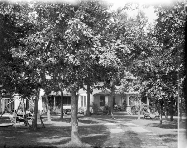 View across lawn of the main building of the Birchcliff Hotel and the tree-lined drive. Vacationers are relaxing on the porch and the lawn.