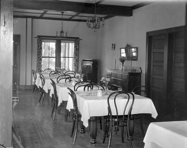 Dining room set with table settings. A mirror and a sideboard are next to the double doors on the right, and a Victrola is in the corner next to two windows. Chandeliers hang from the ceiling.