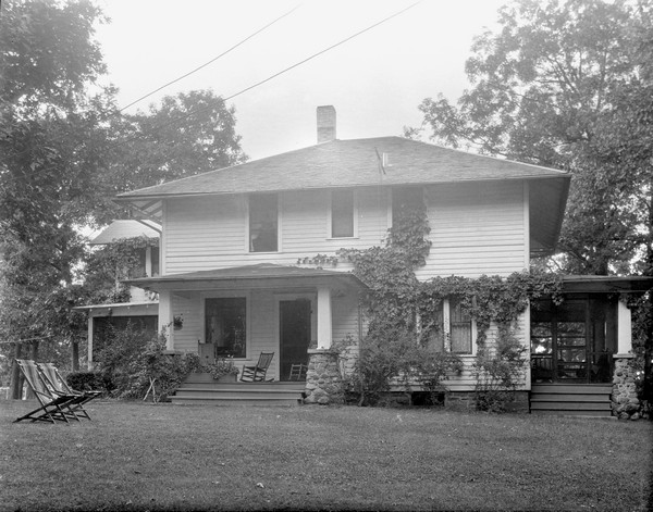 Exterior view across lawn of the main building of the Birchcliff Hotel. A porch is in the front, and a screened-in porch is on the right side. Chairs are on the lawn, and rocking chairs are on the front porch.