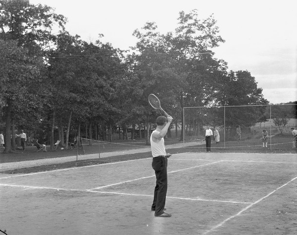 Two men playing tennis on the Birchcliff Hotel tennis court. Another group of guests are playing croquet on the lawn in the background. People relax on chairs under trees on the left.