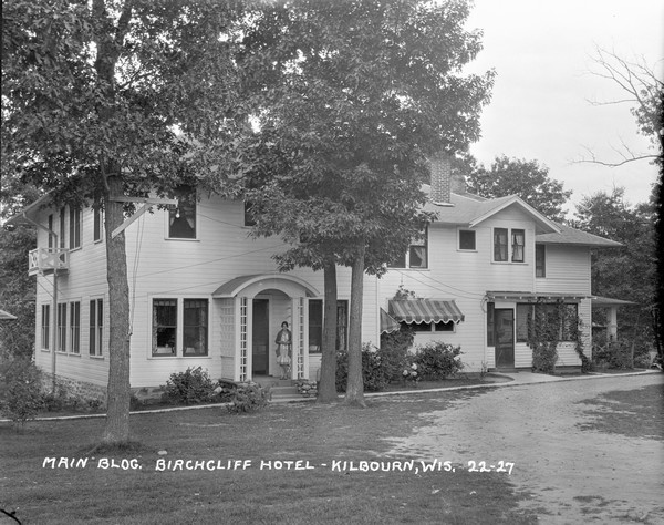 View across lawn of the main building, with a woman is standing at the entrance to the dining room of the Birchcliff Hotel.