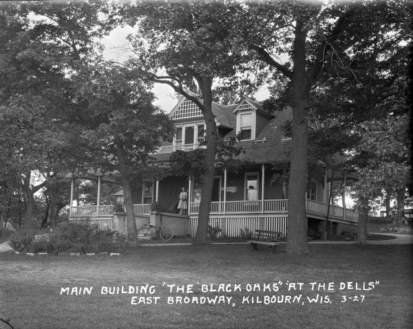 Three-quarter view of the main building. A woman us standing on the porch holding a broom, and a bicycle is parked at the bottom of the stairs. Tennis rackets are hanging on the wall next to the front door. Above the porch is a balcony.