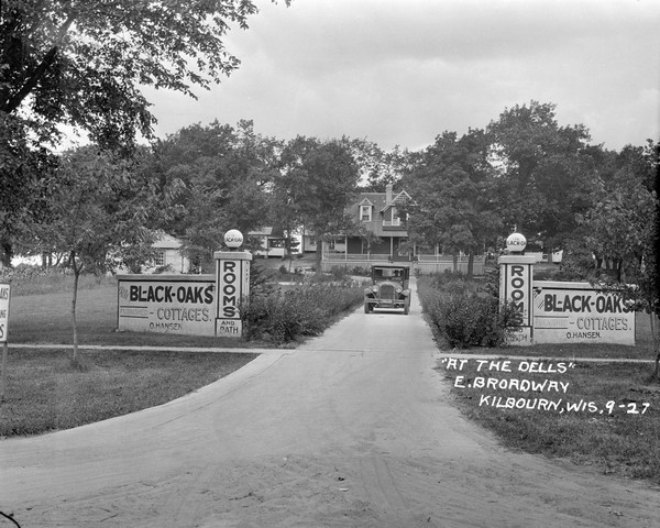 Front entrance to the Black Oaks resort. A man is driving an automobile down the drive near two stone walls with columns framing the entrance to the driveway. They read: "The Black-Oaks." The main building is in the far background, flanked by cottages.