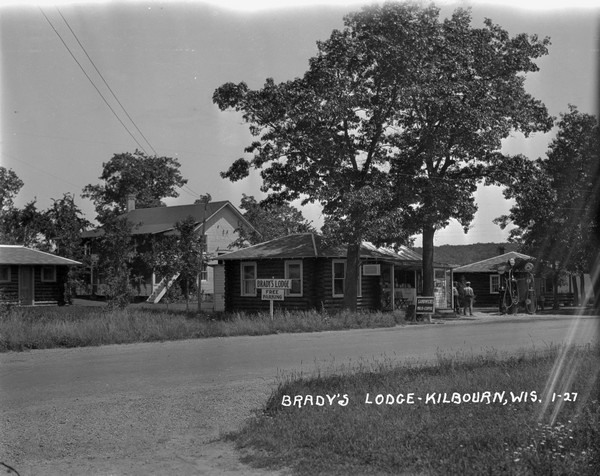 View across road towards the office, filling station and cabin at Brady's Lodge. Two men stand near the gas pumps in front of a porch of an open counter selling food. There are signs for "Rooms," "Free Parking," and "Sandwiches."