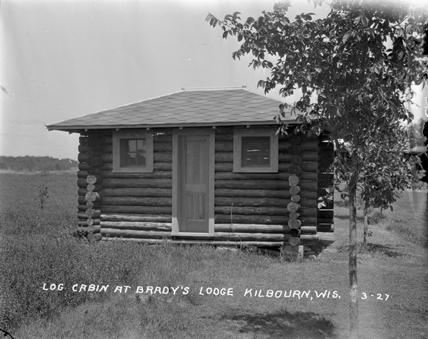 One-room log cabin at Brady's Lodge in a open are with tall grass. Two young trees are along the cabin on the right.