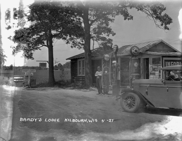 Filling station and office at Brady's Lodge. An attendant is standing by the gas pump. There is a truck parked on the right. Signs on the left read: "Sandwiches," "Free Parking," "Ice Cream" and "Rooms."