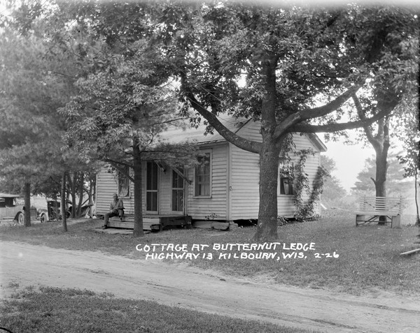 Guest cottage with two doors in front, and a man sitting on the stoop. There are automobiles parked on the left.
