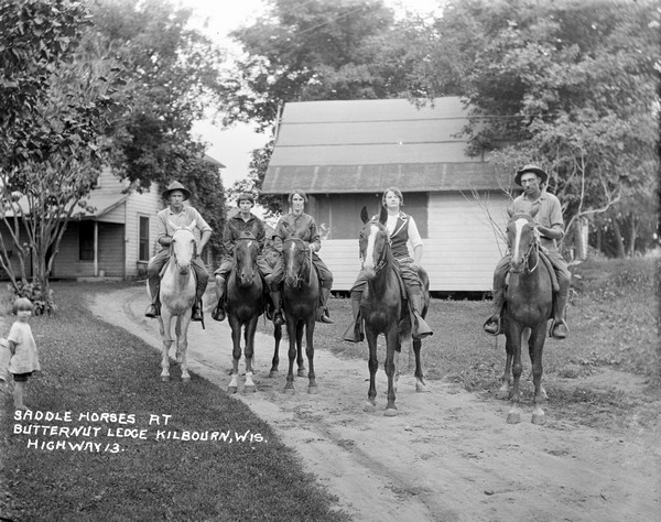 Group portrait of five riders on horses posing on the road in front of two guest cottages. A young girl is standing on the left.
