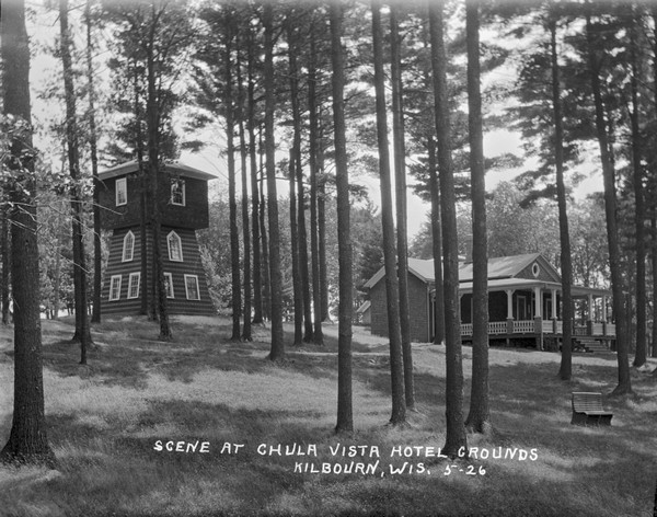 View of grounds with a large guest cottage with large front porch on the right. On the left is a three-story structure shaped like a tower.