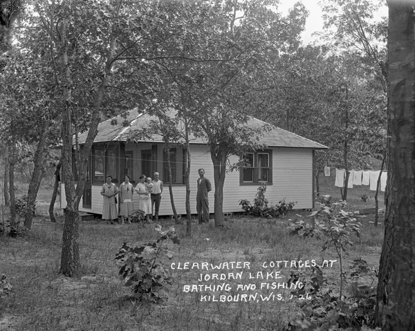 Group of people posing in front of the screened-in porch of a guest cottage. A man stands on the right smoking a pipe, and one of the women is holding a baby. Laundry is drying on a clothesline in back on the right       .