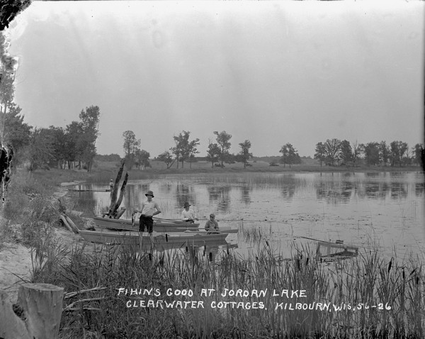 Men and boys fishing at Jordan Lake. One man is standing next to a boat pulled up at the shore and is holding up his catch of the day. There are cattails along the shoreline in the foreground. In the background automobiles are in a field.