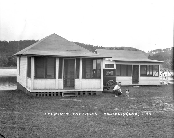 Two Colburn Cottages on a shoreline of the Wisconsin River. In the yard a man is posing in front of an automobile near a dog standing on its haunches. There is a lawn swing on the far right.