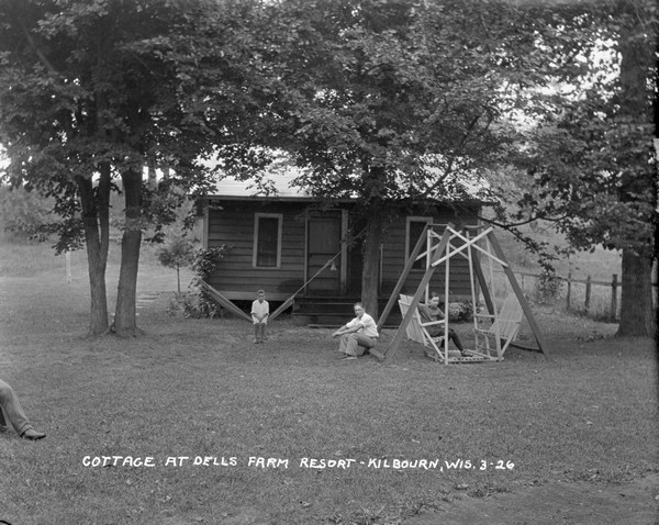 Small cottage with guests sitting in the yard. A young boy sits in a hammock. Two other boys sit on a lawn swing. The leg of another person is on the left in the foreground.