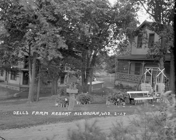 View across road of a large cottage with a front porch on the right, with two people sitting in a lawn chair in front. Down the hill on the left behind trees is a two-story building with a wrap-around porch and a balcony. In the far background two people are sitting in a yard.