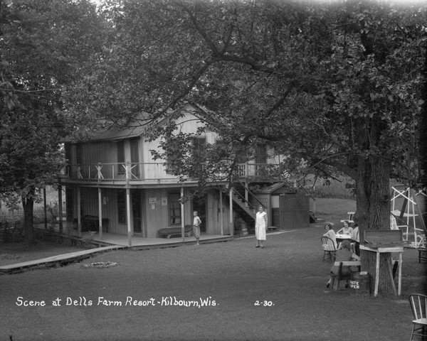Dells Farm Resort with an elevated view of a multi-room cottage with an outhouse located under the balcony on the right. Two women sit around a table in the yard, and a young boy and another woman stand nearby. A woman sits near a tree which has a table holding a croquet set. There is a lawn swing in the background on the right.