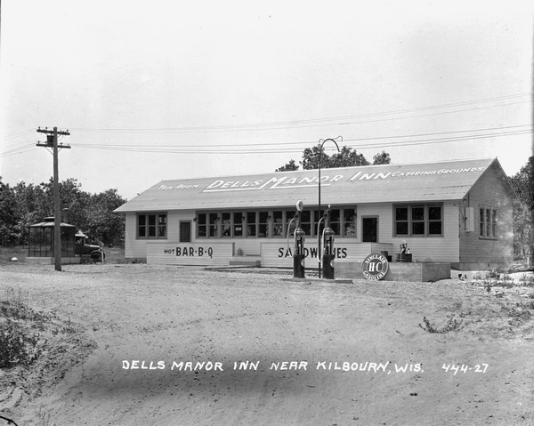 Main building for Dells Manor Inn, Tea Room and Camping Grounds. There are signs for "Hot BBQ" and "Sandwiches," on the side of the building and on the roof. In front are gas pumps with Sinclair gasoline. There is a truck parked on the left.