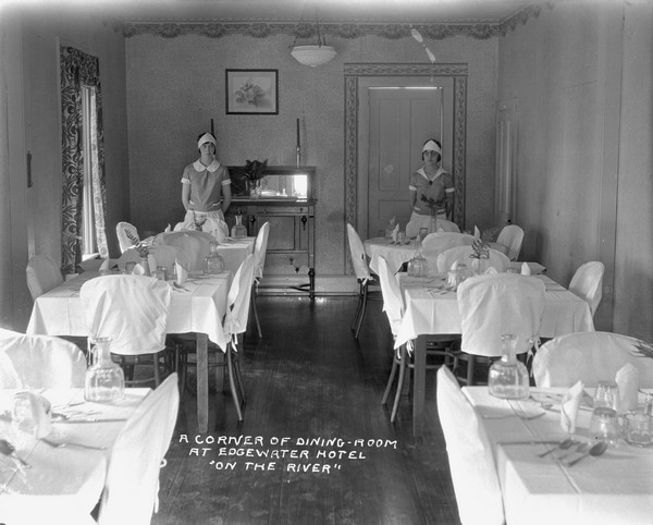 Two waitresses are standing at the back of the dining room at the Edgewater Hotel. All the tables are set with silverware and glasses.