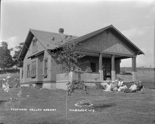 Three-quarter view of Edgewood Valley Resort, a stone dwelling with guest rooms. A group of people is sitting in the lawn in front of the porch.