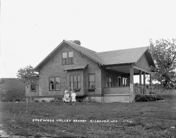 Tree-quarter view of Edgewood Valley Resort, a stone dwelling with guest rooms. A woman and three girls are standing in the yard.