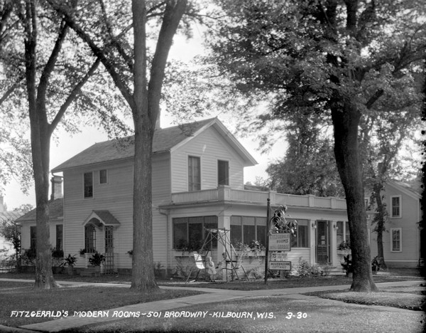 Three-quarter view of Fitzgerald's Modern Rooms, which has a porch and a balcony. Signs in front advertise rooms, bath and garage for 75 cents per person, per night. Two children are on the lawn swing next to the porch.