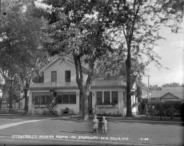 Exterior view of Fitzgerald's Modern Rooms, which is a private home with rooms for tourists, with a screened-in porch, and a balcony. Two children are standing in the lawn, a boy stands on the right near a lawn swing.