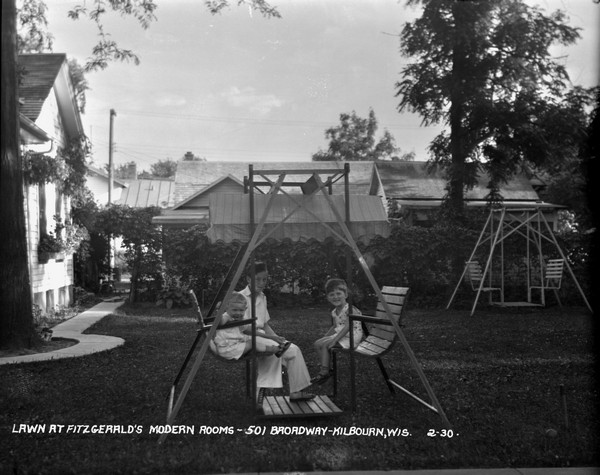 Side lawn of Fitzgerald's Modern Rooms. Children are sitting in the lawn swing in the foreground, and another lawn swing is in the background next to the garden.