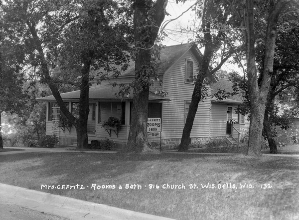 Exterior view of a home that provides rooms and baths for tourists for 75 cents and $1. There is a porch in the front and and in the back. Trees are in the lawn.