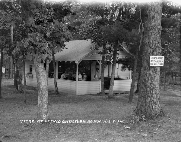 View across lawn of people in the Glenco Cottages store with an open dining area on the porch. A sign on the tree in front reads: "Please do not take or destroy pine trees or ferns."
