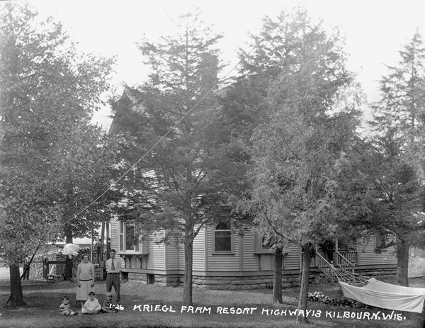 View of the main house at Kriegl Farm Resort. There is a family of five posed on the lawn. A hammock is tied to the trees on the right. More people stand on a porch in the back of the house near a large stack of fuelwood.