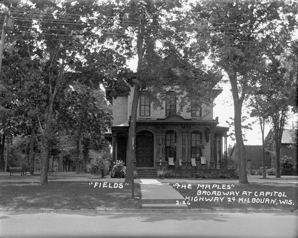 View from street of a large brick house with an arched entrance and wrap-around columned porch. There are benches on the lawn along the side of the house on the left.