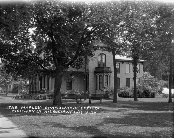 Side view from sidewalk of "The Maples," a large brick house with arched windows and a columned porch.