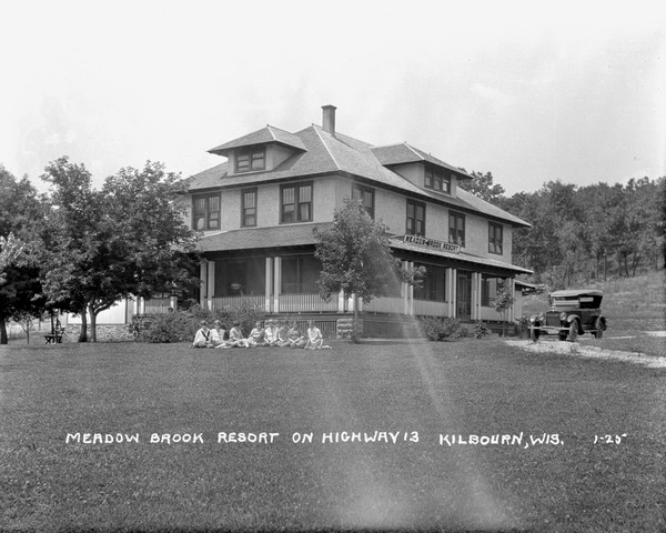 View across lawn of the main building of Meadow Brook Resort. An automobile is parked in the drive and a group of young women are posed sitting in front of the porch on the grass. There is a hill in the background on the right, and a bench and a hand-pump on the left among trees.