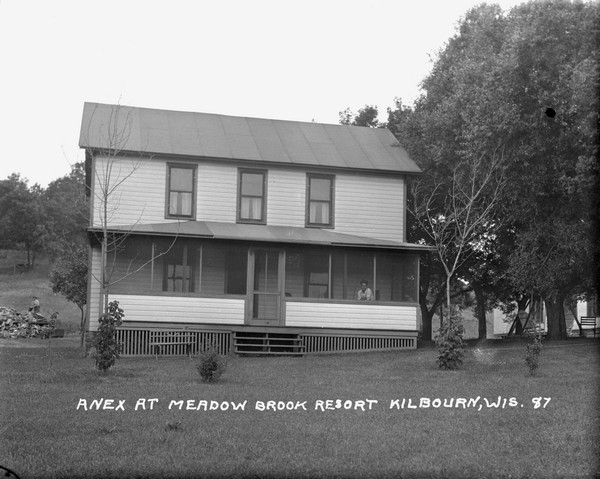 The Annex at Meadow Brook Resort, a two-story building with a screened-in porch. A man is sitting on the porch. A man is standing near a large wood pile in the background on the left. In the side yard on the right is a lawn swing and chairs under the trees.