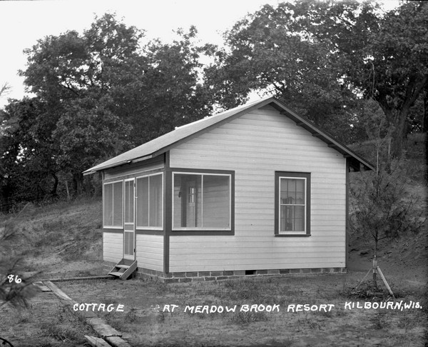 Small cottage with a screened-in porch. There are boards across the dirt yard in the foreground that lead to the door of the porch. A new tree in the yard has supports at the base.