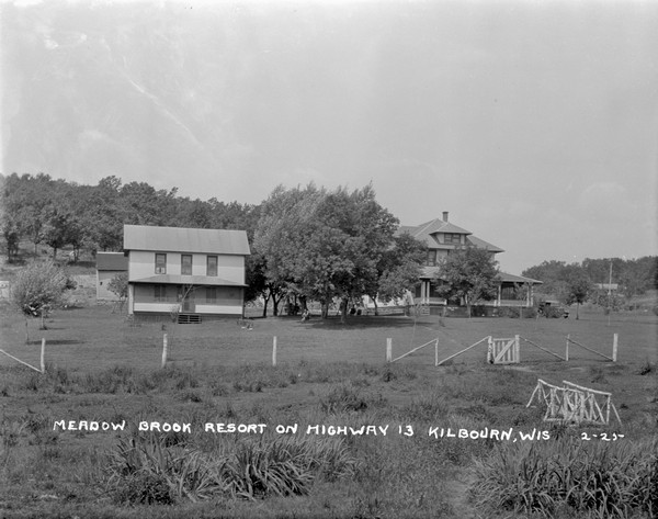 Exterior view of Meadow Brook Resort. The main building is on the right, and the annex is on the left. People are relaxing on the lawn under the trees. There is a fence at the property line, and a foot bridge in the foreground.