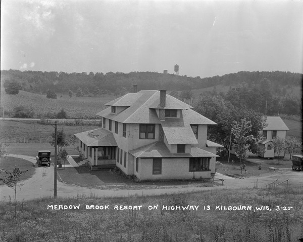 Elevated view from hill of Meadow Brook Resort. The main building is on the left and the annex is on the right. There is an automobile parked in the driveway and another is parked near the annex. A water tower is in the distance on the tree-lined hills.